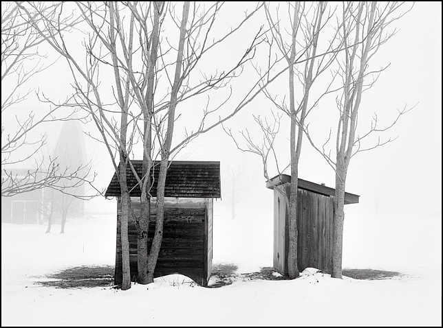 An outhouse and a small shed sit side by side among a group of trees on a farm in southwest Allen County, Indiana. The outhouse and shed are veiled in heavy fog and the ground is covered in snow. A barn is visible through the haze in the background.