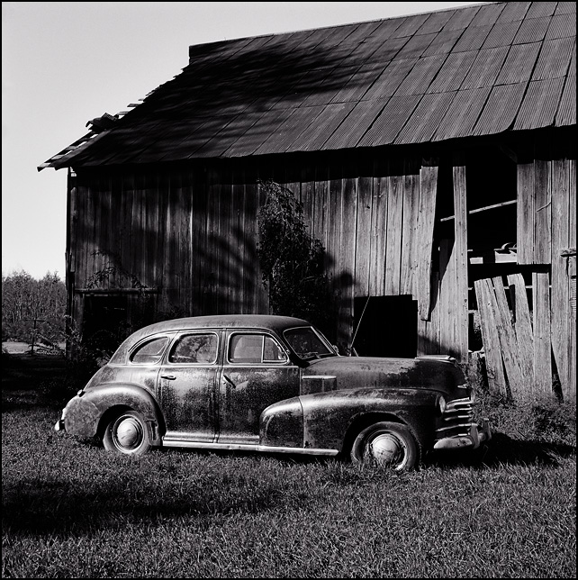 A black 1946 Chevrolet sedan in front of an old barn on a farm in Allen County, Indiana. The shadow of the windmill falls on the barn behind the car.