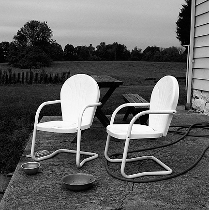A pair of glowing white metal patio chairs behind a farmhouse at sunset in Allen County, Indiana. Dog food bowls, an old license plate, and a picnic table sit on the patio around the chairs.